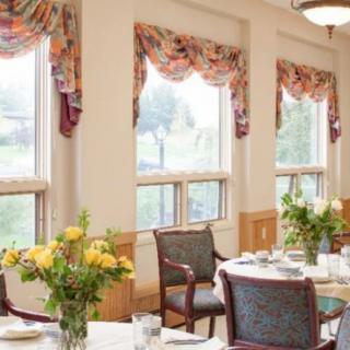 A well-lit dining room with tables set for a meal, featuring decorative curtains and floral arrangements.