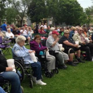 A group of people gathered outdoors, seated on chairs and in wheelchairs, enjoying an event in a park.