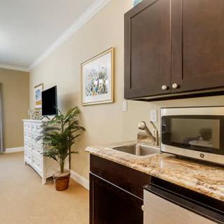 A cozy kitchenette area with a microwave, sink, dark cabinets, and a small seating area visible in the background alongside a television and dresser.