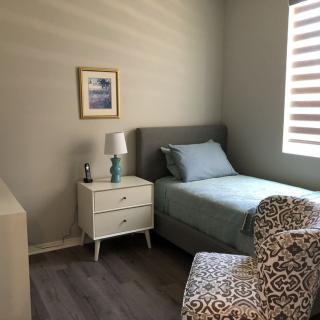 A minimalist bedroom featuring a single bed, nightstand with lamp, patterned chair, and framed artwork on the wall.