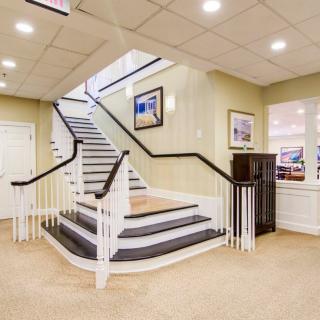 A well-lit lobby featuring a grand staircase with white railings and framed artwork on the walls.