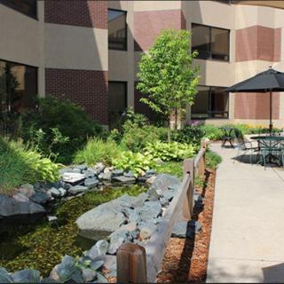 A serene courtyard with a pond, lush greenery, and outdoor seating surrounded by a building's exterior.