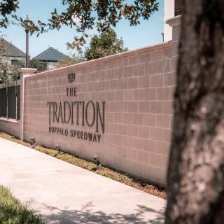 A sign for 'The Tradition' apartment complex displayed on a brick wall along a shaded pathway.