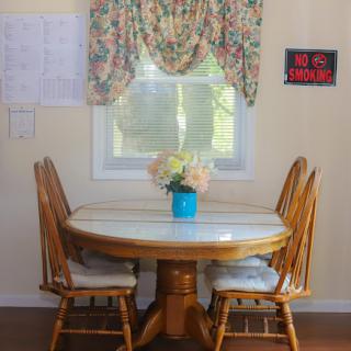 A cozy kitchen space featuring a round dining table surrounded by wooden chairs, adorned with floral curtains and a vibrant flower vase.