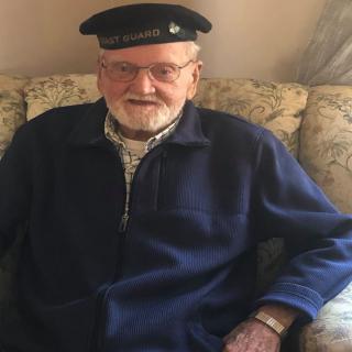 An elderly man wearing a hat and glasses is seated indoors, smiling.