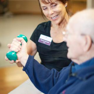 A caregiver assists an elderly person with exercise using a dumbbell.