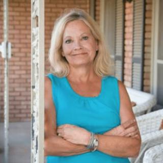 A smiling woman with blonde hair stands on a porch, wearing a blue shirt with her arms crossed.