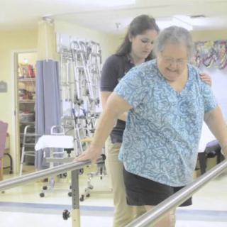 An elderly woman receives assistance from a healthcare professional during a physical therapy session.