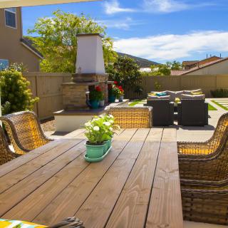 A cozy backyard patio with a wooden dining table, chairs, and an outdoor fireplace under a bright blue sky.