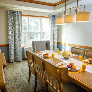 A well-lit dining room featuring a wooden table set with breakfast, surrounded by chairs and accented with elegant curtains.