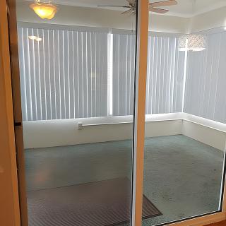 A view of an empty room with large windows and vertical blinds, seen through a glass door.