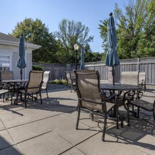 A sunny outdoor patio area with several tables, chairs, and umbrellas surrounded by a fence and trees.