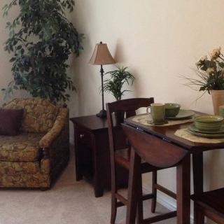 A cozy living room with a dining area, featuring an armchair, side table with a lamp, and a small dining table set with green dishes.
