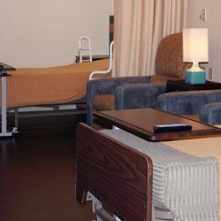 A view of a patient room in a hospital, featuring two beds, a lamp, and seating.