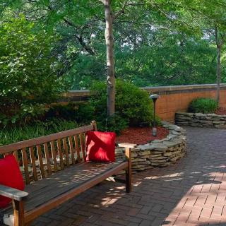 A peaceful garden setting with a wooden bench adorned with red pillows, surrounded by lush greenery and a stone-lined pathway.