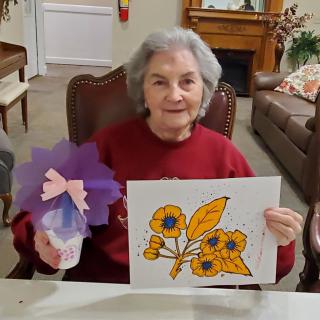 An elderly woman proudly displays her colorful artwork and flower craft.