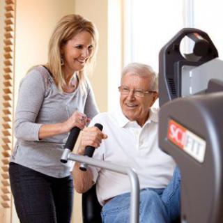 A caregiver assists an elderly man using exercise equipment.
