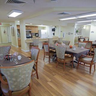 A well-lit dining area with several tables and chairs, featuring wood flooring and an adjacent kitchen.