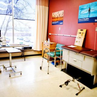 A well-lit medical examination room with various mobility aids like a wheelchair and walker, along with medical equipment and a desk.