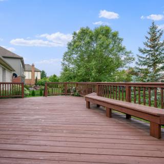 A spacious wooden deck with built-in benches, surrounded by greenery and neighboring houses.