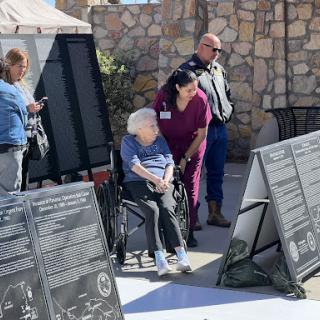 An elderly individual in a wheelchair observes informational displays at an outdoor event, assisted by a caregiver.