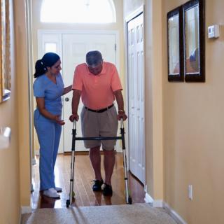 A nurse assists an elderly man using a walker in a home setting.