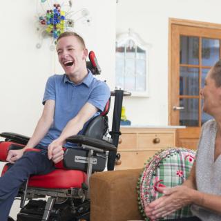 A young person in a wheelchair is smiling and talking with another individual indoors.