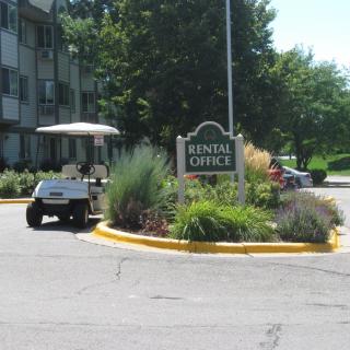 A rental office sign located near an apartment complex, with a golf cart parked nearby and landscaping in the surroundings.