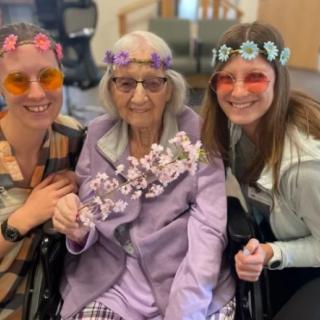 Three women, one elderly and in a wheelchair, wearing flower crowns and smiling.