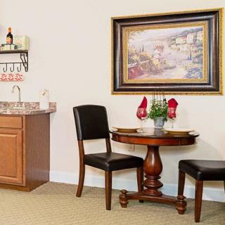 A cozy dining area with a round table set for two, accompanied by a painting and a small kitchenette with a wine rack.