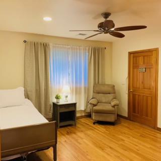 A cozy bedroom with wooden flooring, featuring a bed, armchair, ceiling fan, and a side table with a lamp.