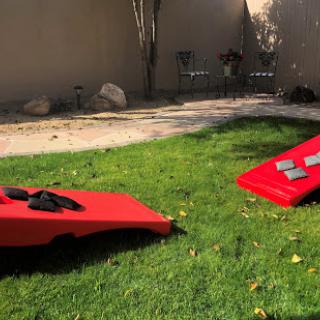 A sunny backyard setup featuring two red cornhole boards on a grassy lawn.