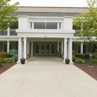 A modern building entrance with white columns and a paved walkway, surrounded by greenery.