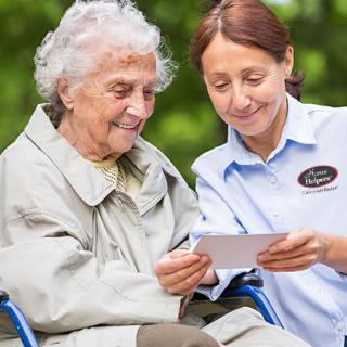 A caregiver and an elderly woman share a moment outdoors, smiling as they look at a photo together.