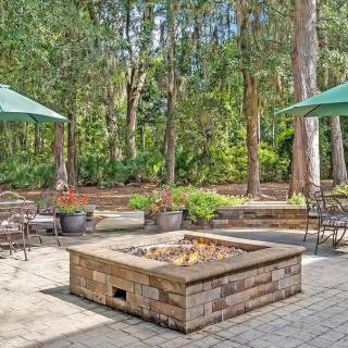 A serene outdoor patio area featuring a central fire pit, surrounded by seating and large umbrellas, nestled in a forested setting.