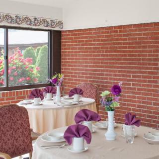 A cozy dining room with tables set elegantly with purple napkins and floral centerpieces, next to a large window showcasing a garden view.