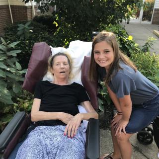 A young girl poses next to an elderly woman in a wheelchair, set in a garden.