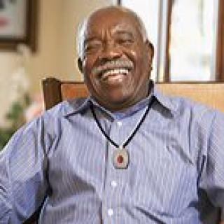 An elderly man smiling while sitting indoors, wearing a striped shirt and necklace.