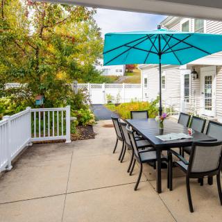 A backyard patio with a dining set and blue umbrella, surrounded by greenery.