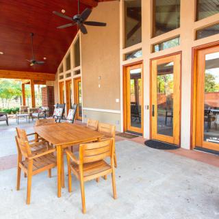 An outdoor patio area with wooden dining furniture and ceiling fans, adjacent to a building with large glass doors.