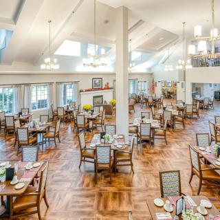 A spacious and elegantly decorated dining area with multiple tables and chairs, featuring a wooden floor and bright lighting from chandeliers.