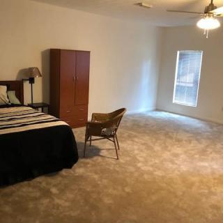 A spacious bedroom with a bed, wardrobe, and chair on a carpeted floor, featuring a window and ceiling fan.