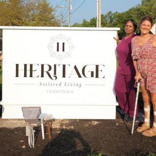 Two people standing next to a Heritage Assisted Living sign outdoors.