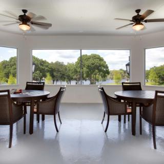 A bright dining area with multiple tables and chairs, featuring large windows and ceiling fans.