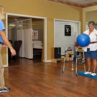 An elderly person engages in a physical therapy session using a fitness ball, assisted by a therapist indoors.