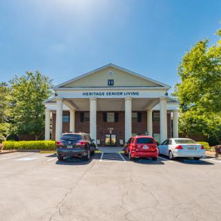 A front view of a building with large columns, surrounded by trees, and several cars parked in the lot on a sunny day.