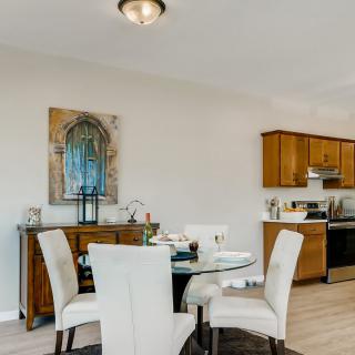 A modern dining space with a round glass table and upholstered chairs, adjacent to a kitchen area with wooden cabinets.