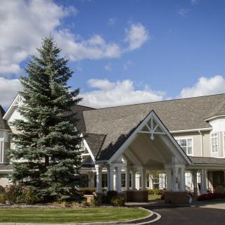 A large residential building with a steep roof and a tall tree in front, set against a backdrop of blue sky and clouds.