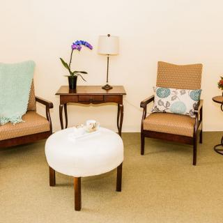 A cozy living room setup featuring two armchairs with cushions, a small side table with a lamp and orchid plant, and a round ottoman in the center.