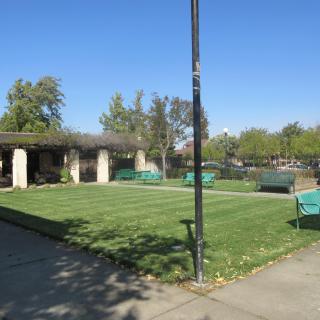 A sunny day at a park featuring a grassy area, benches, and a pavilion surrounded by trees.